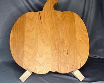 May include: A pumpkin-shaped wooden cutting board with a light brown finish. The board is supported by two small wooden stands. The board has a stem and a curved shape, and is ideal for fall-themed kitchen decor.