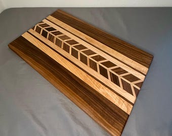 May include: Rectangular wooden cutting board with a decorative arrow design. The board features alternating stripes of dark and light wood, with a central arrow pattern. The wood tones range from light tan to dark brown.