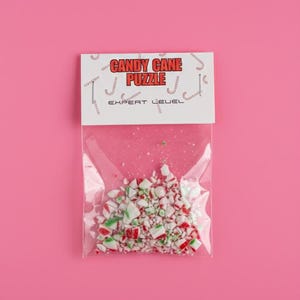 Puede incluir: Una bolsa de plástico transparente llena de caramelos de bastón triturados, con trozos rojos, blancos y verdes. La bolsa tiene un encabezado blanco con el texto "CANDY CANE PUZZLE" en rojo y negro, y "EXPERT LEVEL" debajo.