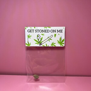 May include: A clear plastic bag with a small, green object inside, and a white label that says "GET STONED ON ME". The label has a drawing of a lit joint and cannabis leaves. The background is a solid pink colour.