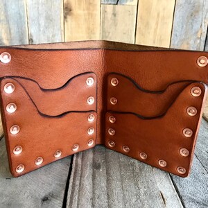 May include: Open brown leather wallet with multiple card slots. The wallet is secured with copper rivets along the edges. The interior features additional pockets for organization. The wallet is displayed open, showcasing its design and construction.