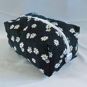 May include: Black zippered pouch with a white daisy print on a black background. The pouch has a white zipper and a white pull tab.
