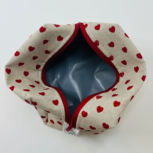 Puede incluir: Una bolsa de cremallera beige con un patr&oacute;n de corazones rojos. La bolsa est&aacute; abierta y el interior est&aacute; forrado con un tejido gris.