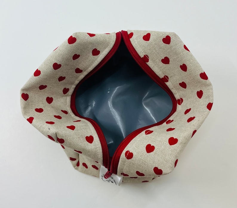 Puede incluir: Una bolsa de cremallera beige con un patr&oacute;n de corazones rojos. La bolsa est&aacute; abierta y el interior est&aacute; forrado con un tejido gris.