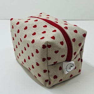 Puede incluir: Una bolsa de cremallera beige con un patr&oacute;n de corazones rojos y una etiqueta blanca que dice "The Crafty Craft Company".