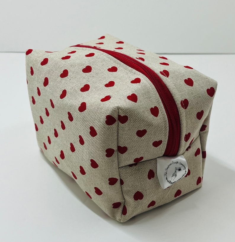 Puede incluir: Una bolsa de cremallera beige con un patr&oacute;n de corazones rojos y una etiqueta blanca que dice "The Crafty Craft Company".