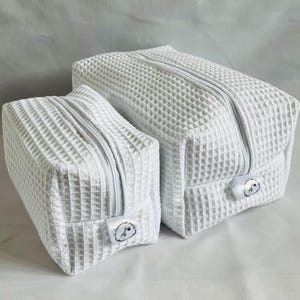 Può includere: Due pochette con cerniera bianche a trama a nido d'ape. La pochette più grande è a destra e quella più piccola a sinistra. Entrambe le pochette hanno una cerniera bianca e un piccolo logo bianco.
