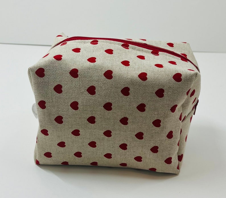 Puede incluir: Una bolsa de cremallera beige con un patr&oacute;n de corazones rojos.
