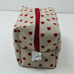 Puede incluir: Una bolsa de cremallera beige con un patr&oacute;n de corazones rojos. La bolsa tiene una etiqueta blanca con el texto "The Crafty Corner Company".
