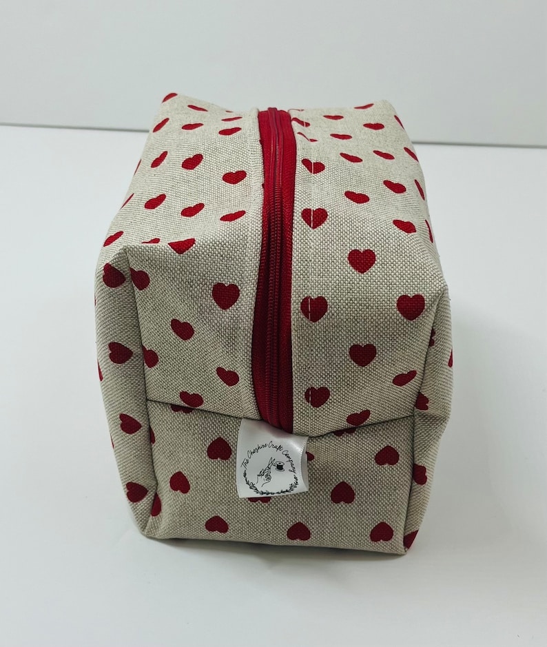 Puede incluir: Una bolsa de cremallera beige con un patr&oacute;n de corazones rojos. La bolsa tiene una etiqueta blanca con el texto "The Crafty Corner Company".