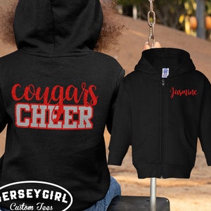 May include: Black zippered hoodie with red glitter text that says "cougars cheer" on the back and "Jasmine" on the front.  The hoodie is shown on a child wearing blue jeans.