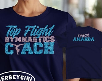 May include: Navy blue t-shirt with glitter text that reads "Top Flight Gymnastics Coach" with a silhouette of a gymnast. The back of the shirt has the text "Coach Amanda" in glitter.