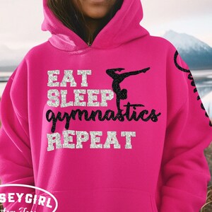 May include: Pink hoodie with a black silhouette of a gymnast doing a handstand and the text "Eat Sleep Gymnastics Repeat" in silver glitter.