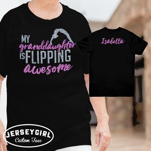 Puede incluir: Camiseta negra con el texto "MY granddaughter IS FLIPPING Awesome" en purpurina plateada y rosa. Una segunda camiseta negra muestra el nombre "Isabella" en rosa. Las camisetas son de Jersey Girl Custom Tees.