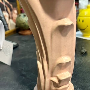 May include: A pale pink ceramic vase with a unique, abstract design. The vase has a wide opening and a narrow base, and it is decorated with a series of raised lines and curves.
