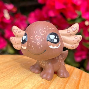 May include: A hand-painted brown axolotl figurine with white hearts and dots. The figurine has large black eyes with white pupils and a small, closed mouth.