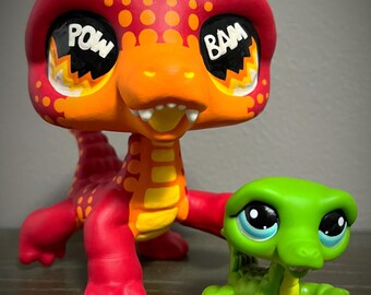 May include: Two colorful, hand-painted reptile figurines. The larger one is red and orange with "POW" and "BAM" in its eyes. The smaller one is green. Both have cartoonish features and are posed as if in action.