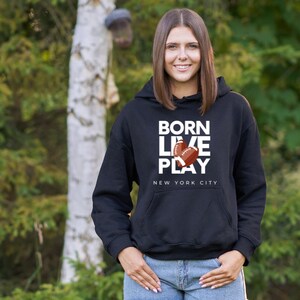 K&ouml;nnte beinhalten: Schwarzer Hoodie mit wei&szlig;em Text, der "Born Live Play" lautet, mit einer Grafik von zwei Fu&szlig;b&auml;llen. Der Text "New York City" befindet sich unter der Grafik.