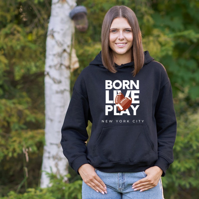 K&ouml;nnte beinhalten: Schwarzer Hoodie mit wei&szlig;em Text, der "Born Live Play" lautet, mit einer Grafik von zwei Fu&szlig;b&auml;llen. Der Text "New York City" befindet sich unter der Grafik.