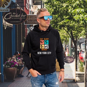 K&ouml;nnte beinhalten: Schwarzer Kapuzenpullover mit einem Grafikdruck einer Person, die Fahrrad f&auml;hrt, und dem Text "Eat Sleep Repeat New York City".