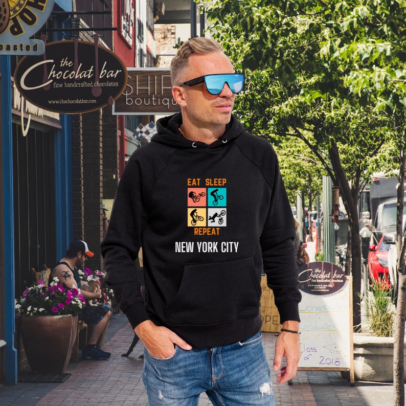 K&ouml;nnte beinhalten: Schwarzer Kapuzenpullover mit einem Grafikdruck einer Person, die Fahrrad f&auml;hrt, und dem Text "Eat Sleep Repeat New York City".