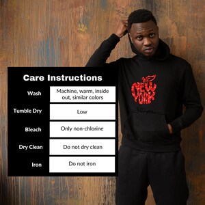 K&ouml;nnte beinhalten: Schwarzer Hoodie mit einem roten Apfel-Motiv, auf dem "New York" und "Ich liebe dich" steht. Pflegehinweise sind auf wei&szlig;em Hintergrund aufgelistet: Maschinenw&auml;sche warm, auf links, &auml;hnliche Farben. Trocknergeeignet bei niedriger Temperatur. Nur nicht-chlorhaltiges Bleichmittel verwenden. Nicht chemisch reinigen. Nicht b&uuml;geln.