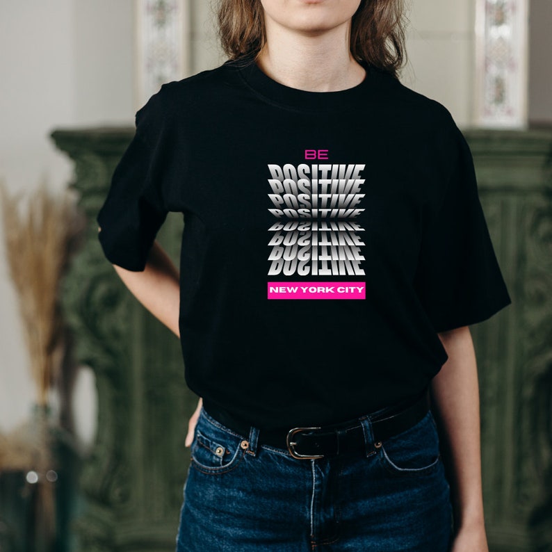 K&ouml;nnte beinhalten: Ein schwarzes T-Shirt mit einer wei&szlig;en Grafik, auf der "BE POSITIVE" in einem sich wiederholenden Muster steht. Der Text ist im 3D-Stil gehalten und leicht angewinkelt. Der Text "NEW YORK CITY" ist in Pink unter der Grafik gedruckt.