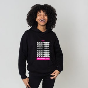 K&ouml;nnte beinhalten: Schwarzer Hoodie mit einem Grafikdruck, der "BE POSITIVE" in einem sich wiederholenden Muster mit immer kleiner werdenden W&ouml;rtern sagt. Der Text "NEW YORK CITY" ist in Pink unter dem Grafikdruck gedruckt.