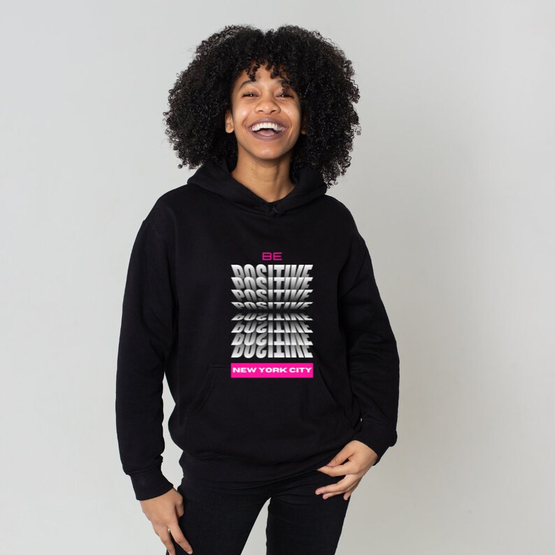 K&ouml;nnte beinhalten: Schwarzer Hoodie mit einem Grafikdruck, der "BE POSITIVE" in einem sich wiederholenden Muster mit immer kleiner werdenden W&ouml;rtern sagt. Der Text "NEW YORK CITY" ist in Pink unter dem Grafikdruck gedruckt.