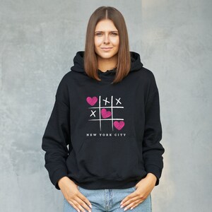 K&ouml;nnte beinhalten: Schwarzer Kapuzenpullover mit einem pinkfarbenen und wei&szlig;en Grafikdruck eines Tic-Tac-Toe-Spielfelds mit Herzen und dem Text "NEW YORK CITY".