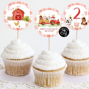May include: Three cupcakes decorated with farm animal themed toppers. The toppers are round and feature a red barn, a red truck with farm animals, and a number 2 with farm animals. The cupcakes are frosted with white frosting and are on a white doily.