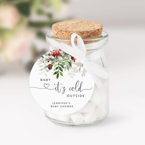 Op de afbeelding: Een klein glazen potje gevuld met witte snoepjes, vastgebonden met een wit lint en een rond etiket waarop staat "Baby, het is koud buiten" met een klein hartje en een groene illustratie. Het etiket vermeldt ook "Jennifer's Baby Shower".