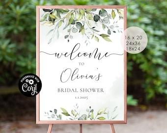 May include: A white welcome sign with a watercolor floral design in shades of green and gray. The sign reads "Welcome to Olivia's Bridal Shower 11.1.2025". The sign is framed in a light brown frame and is displayed on a wooden easel.