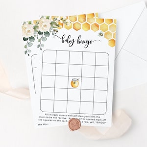 May include: A white baby bingo card with a honeycomb pattern and a bee illustration. The card has a free space in the center and 24 other squares to fill in with gift items. The text "baby bingo" is written in black cursive at the top of the card.