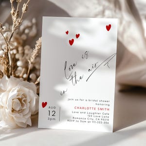 May include: A white invitation card with red heart accents and the phrase "Love is in the air" in elegant script. The card announces a bridal shower for Charlotte Smith, with date, time, and location details.