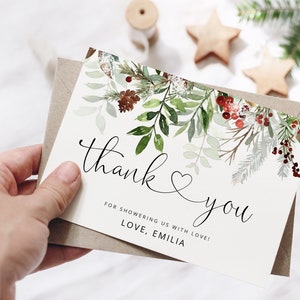 May include: A white thank you card with a watercolor illustration of green leaves, red berries, and pine cones. The text on the card reads "thank you" with a heart symbol and "for showering us with love! Love, Emilia."