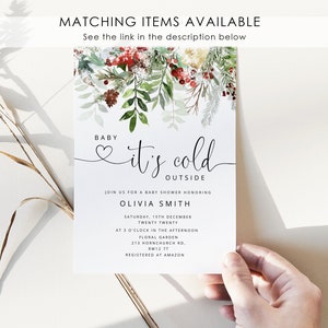 May include: A white baby shower invitation with a watercolor illustration of winter greenery and red berries. The text reads "Baby it's cold outside" and includes details for the event, including the date, time, and location. The invitation is registered at Amazon.