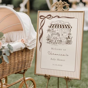 Op de afbeelding: Een rechthoekig bord in een gouden lijst met een bruine lintillustratie, een schets van een café en de tekst: Welcome to Veronica's Baby Shower 11.12.2029. Het bord heeft een beige en wit gestreepte achtergrond en staat op een gouden standaard.