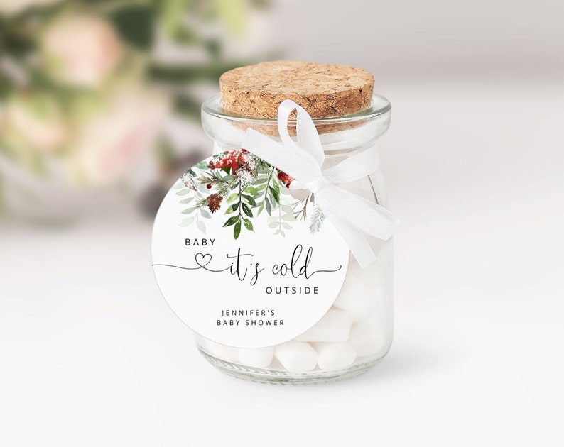 Op de afbeelding: Een klein glazen potje gevuld met witte snoepjes, vastgebonden met een wit lint en een rond etiket waarop staat "Baby, het is koud buiten" met een klein hartje en een groene illustratie. Het etiket vermeldt ook "Jennifer's Baby Shower".