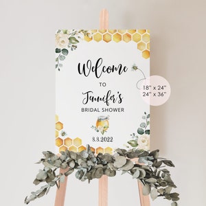 May include: Welcome to Jennifer's Bridal Shower sign with a honey pot and bee design. The sign is decorated with white flowers and green leaves. The sign is available in two sizes: 18" x 24" and 24" x 36".