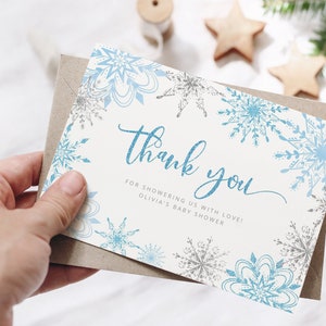 Pu&ograve; includere: Un biglietto di ringraziamento a tema invernale. La carta presenta disegni di fiocchi di neve blu e argento sui bordi e le parole "Thank you" in un carattere blu glitterato. La carta &egrave; in una busta marrone.
