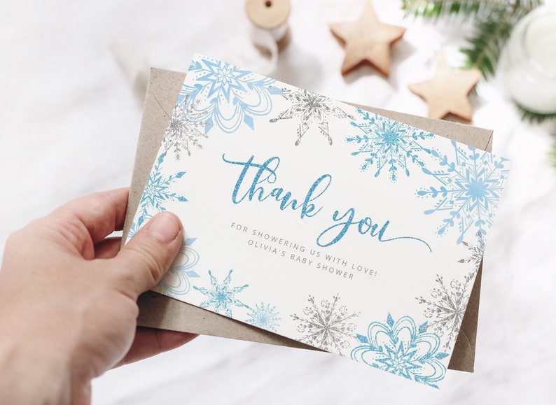 Pu&ograve; includere: Un biglietto di ringraziamento a tema invernale. La carta presenta disegni di fiocchi di neve blu e argento sui bordi e le parole "Thank you" in un carattere blu glitterato. La carta &egrave; in una busta marrone.