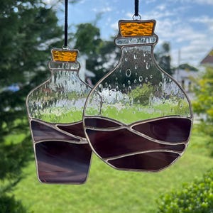 May include: Two stained glass bottle ornaments with cork tops. The bottles are clear with a textured surface and filled with a dark purple liquid. The bottles are hanging from black cords.