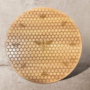 May include: A round, light-colored wooden cutting board with a laser-etched bee and honeycomb design. The board features a large bee in the center, surrounded by smaller bees and a hexagonal pattern. The board is approximately 12 inches in diameter.