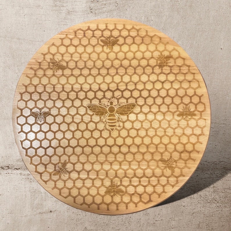 May include: A round, light-colored wooden cutting board with a laser-etched bee and honeycomb design. The board features a large bee in the center, surrounded by smaller bees and a hexagonal pattern. The board is approximately 12 inches in diameter.