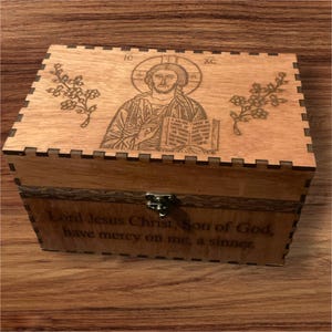May include: A wooden box with a hinged lid. The lid features a laser-engraved image of Jesus Christ holding a book. The inscription "Lord Jesus Christ, Son of God, have mercy on me, a sinner." is engraved on the front of the box.