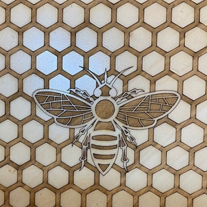 May include: A laser-cut wooden bee design, centered on a honeycomb pattern. The bee is intricately detailed with segmented wings and body, set against a backdrop of hexagonal cells. The wood has a natural, light brown color.