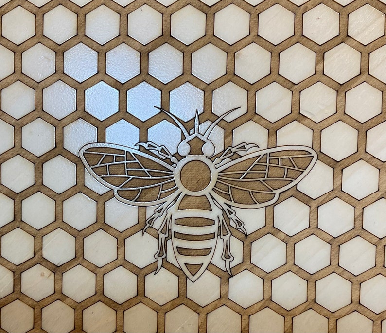 May include: A laser-cut wooden bee design, centered on a honeycomb pattern. The bee is intricately detailed with segmented wings and body, set against a backdrop of hexagonal cells. The wood has a natural, light brown color.