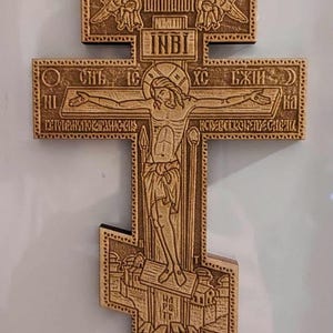May include: A wooden Orthodox cross with intricate carvings. The center features a depiction of Jesus Christ on the cross, with angels and other religious symbols surrounding it. The inscription "INBI" is visible at the top of the cross.