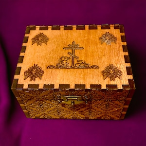 May include: A wooden box with a hinged lid, featuring intricate carvings. The box is light brown with a darker brown border and a gold-colored clasp. The lid is decorated with a cross and angel motifs, and the sides have a floral pattern.